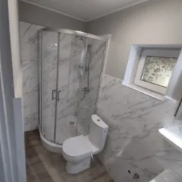 Ukończona łazienka z kabiną prysznicową z zaokrąglonymi drzwiami, toaletą typu kompakt i umywalką, wykończona płytkami imitującymi biały marmur z szarymi żyłami oraz drewnopodobnymi płytkami...