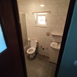 MattBud - Widok na odnowioną łazienkę z kabiną prysznicową, umywalką z szafką i toaletą, sfotografowaną zza uchylonych drzwi z widocznym fragmentem futryny.