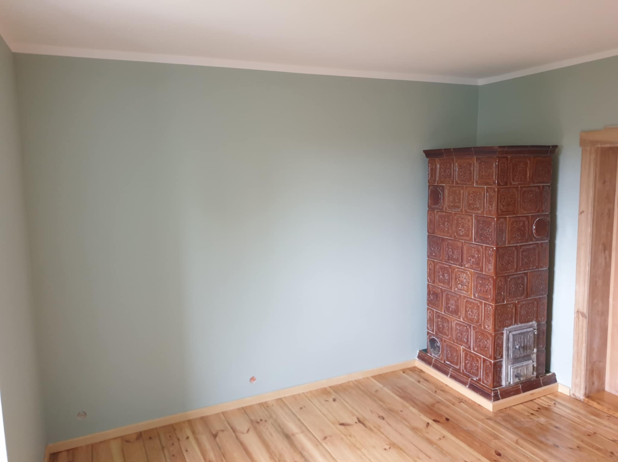 Pomalowane na jasnozielono ściany w pokoju z drewnianą podłogą i piecem kaflowym w rogu, widoczny fragment drewnianych drzwi.