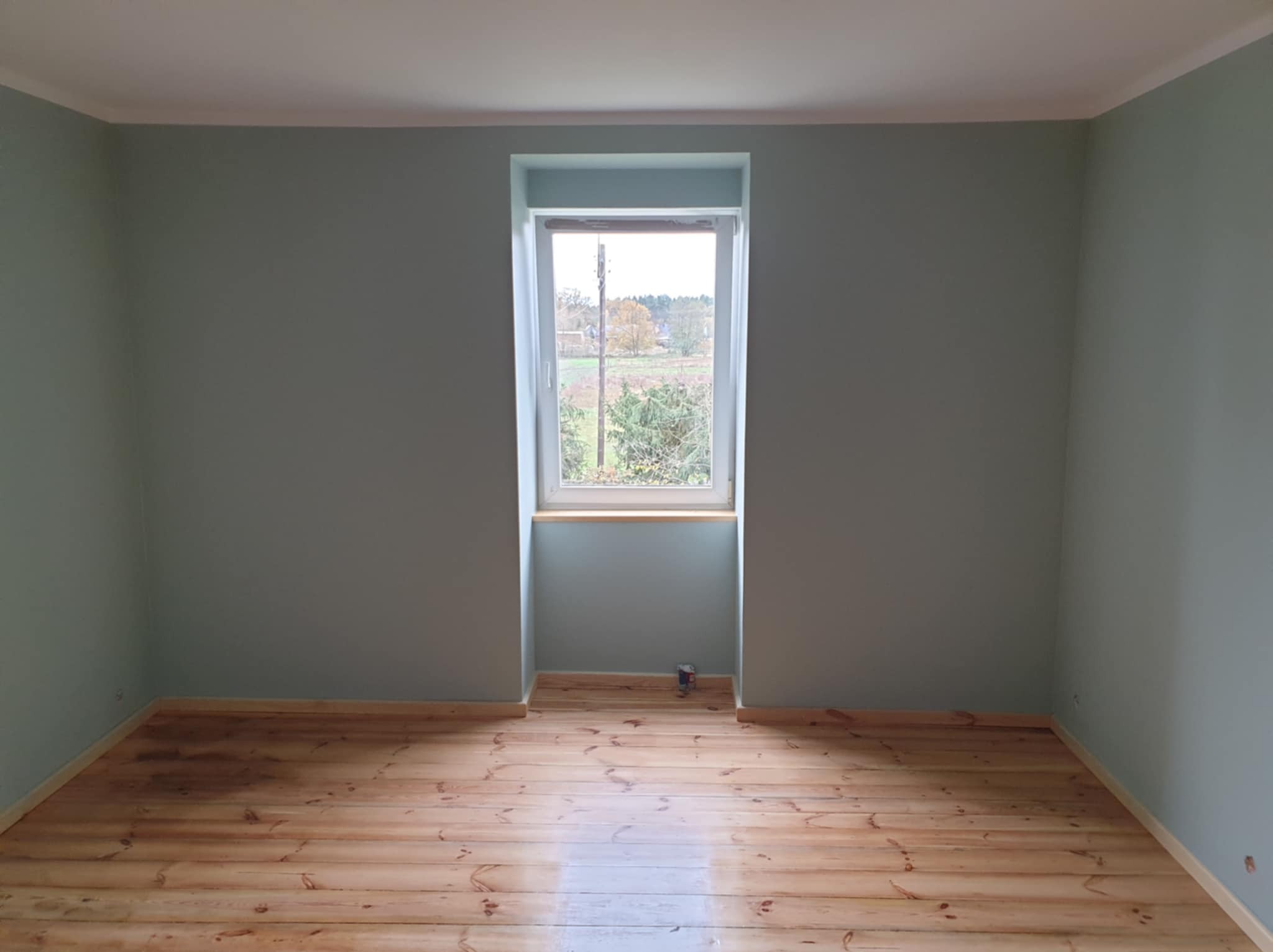 Pomalowane na pastelowo-zielono puste pomieszczenie z drewnianą podłogą i oknem wychodzącym na zewnątrz, widoczna puszka po farbie przy ścianie.