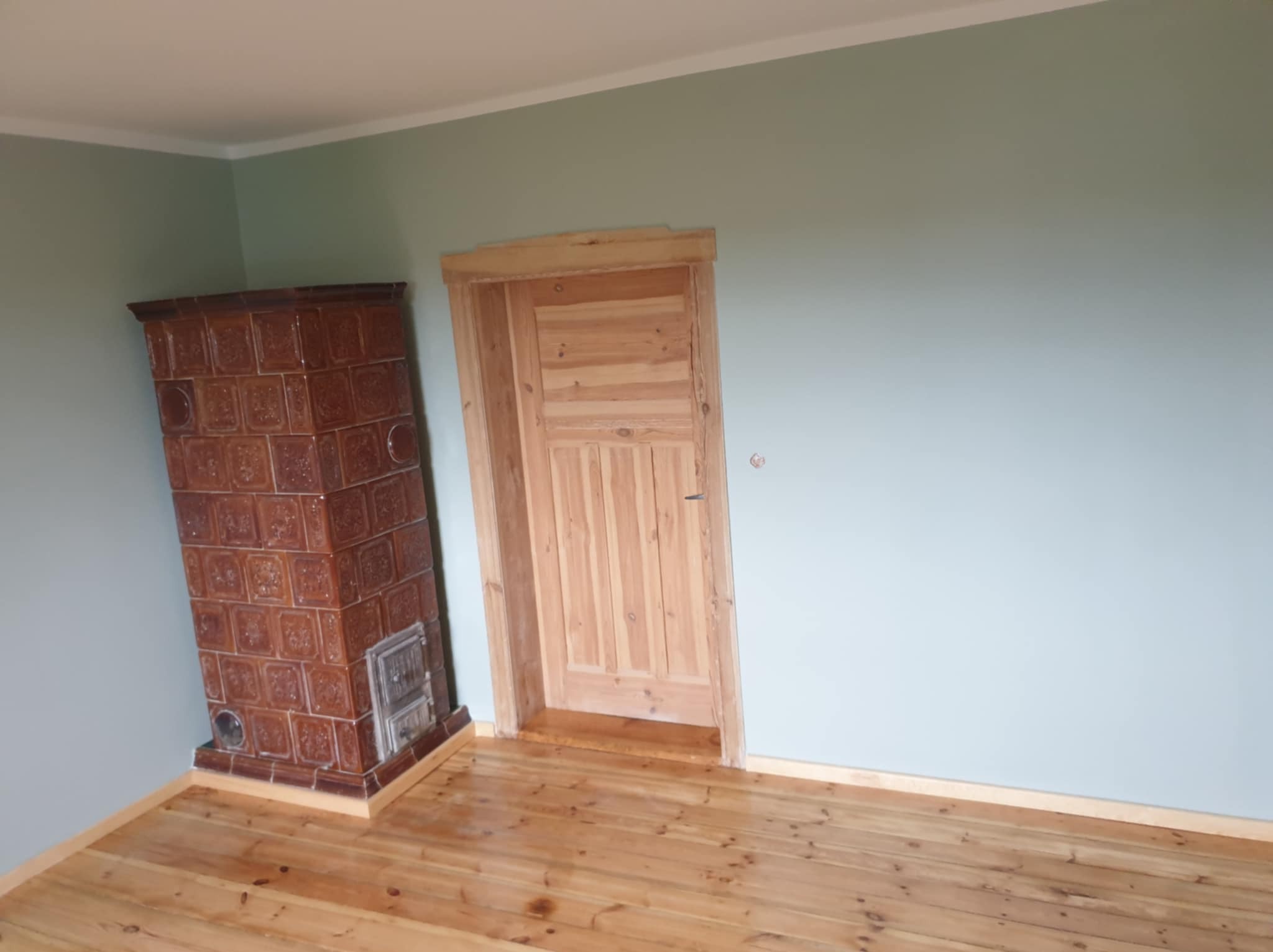 Wnętrze pokoju po remoncie: świeżo pomalowane ściany w odcieniach szarości, nowa drewniana podłoga, piec kaflowy z brązowych płytek oraz surowe drewniane drzwi z ościeżnicą.