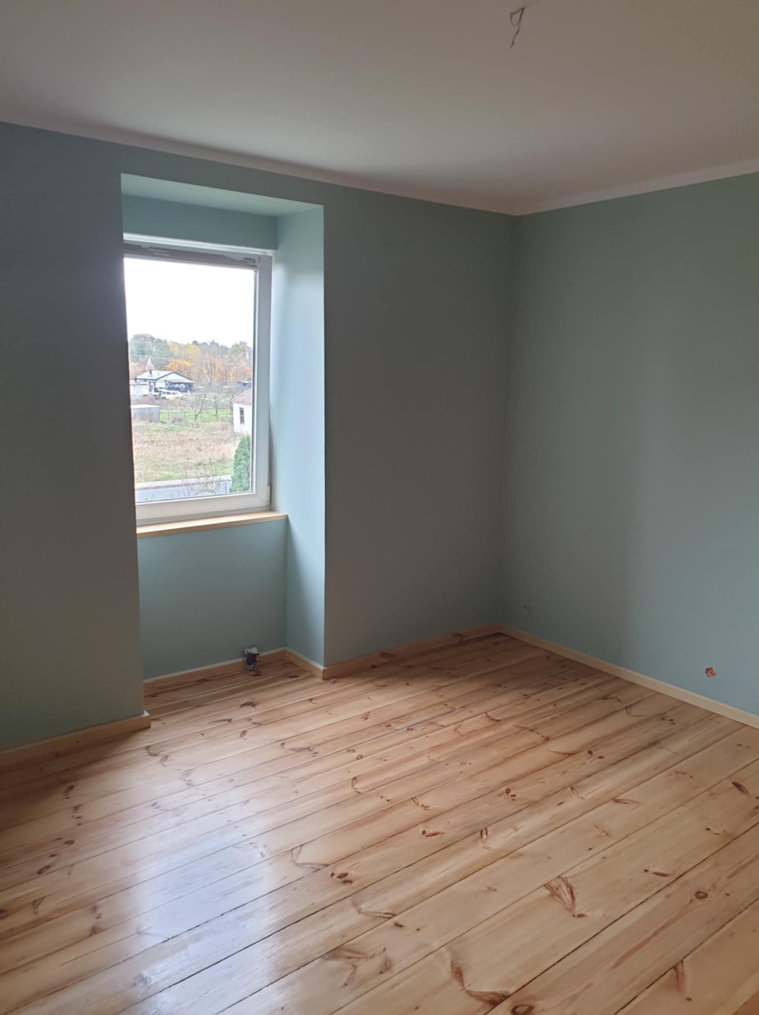 Pomalowane na pastelowo zielono wnętrze pokoju z drewnianą podłogą, widok na zewnątrz przez okno z drewnianym parapetem, w rogu pod oknem stoi puszka z farbą.