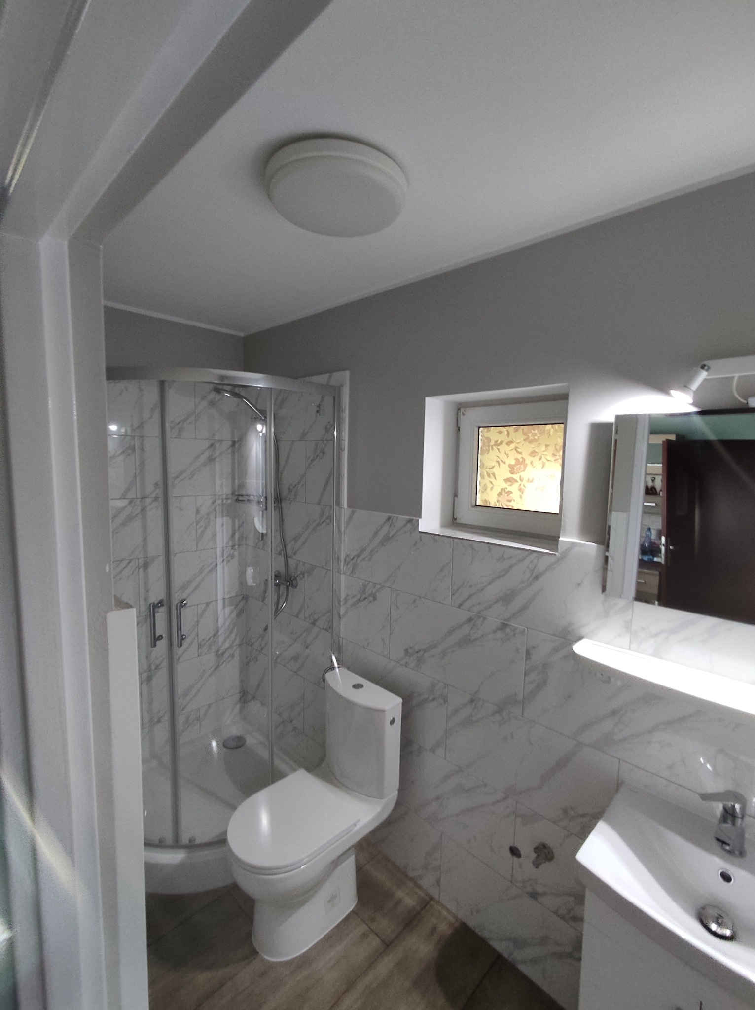 Jasna łazienka po remoncie: kabina prysznicowa z marmurowymi płytkami, biała umywalka z szafką i toaleta typu kompakt. Małe okno z ozdobną folią.