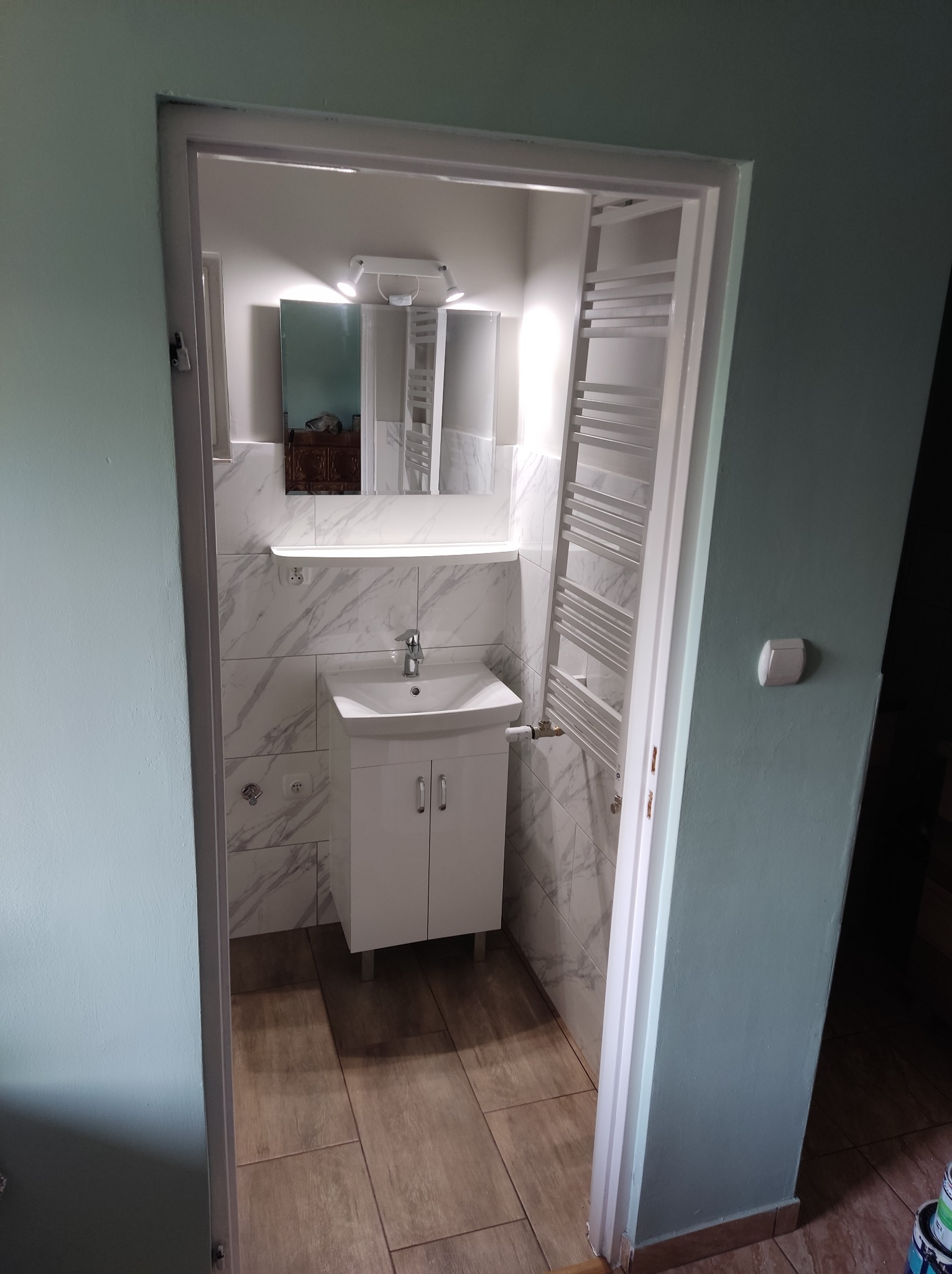 Wykończona łazienka z białymi płytkami imitującymi marmur, białą szafką z umywalką, grzejnikiem drabinkowym i lustrem z oświetleniem, widok z korytarza z pomalowanymi na zielono ścianami.