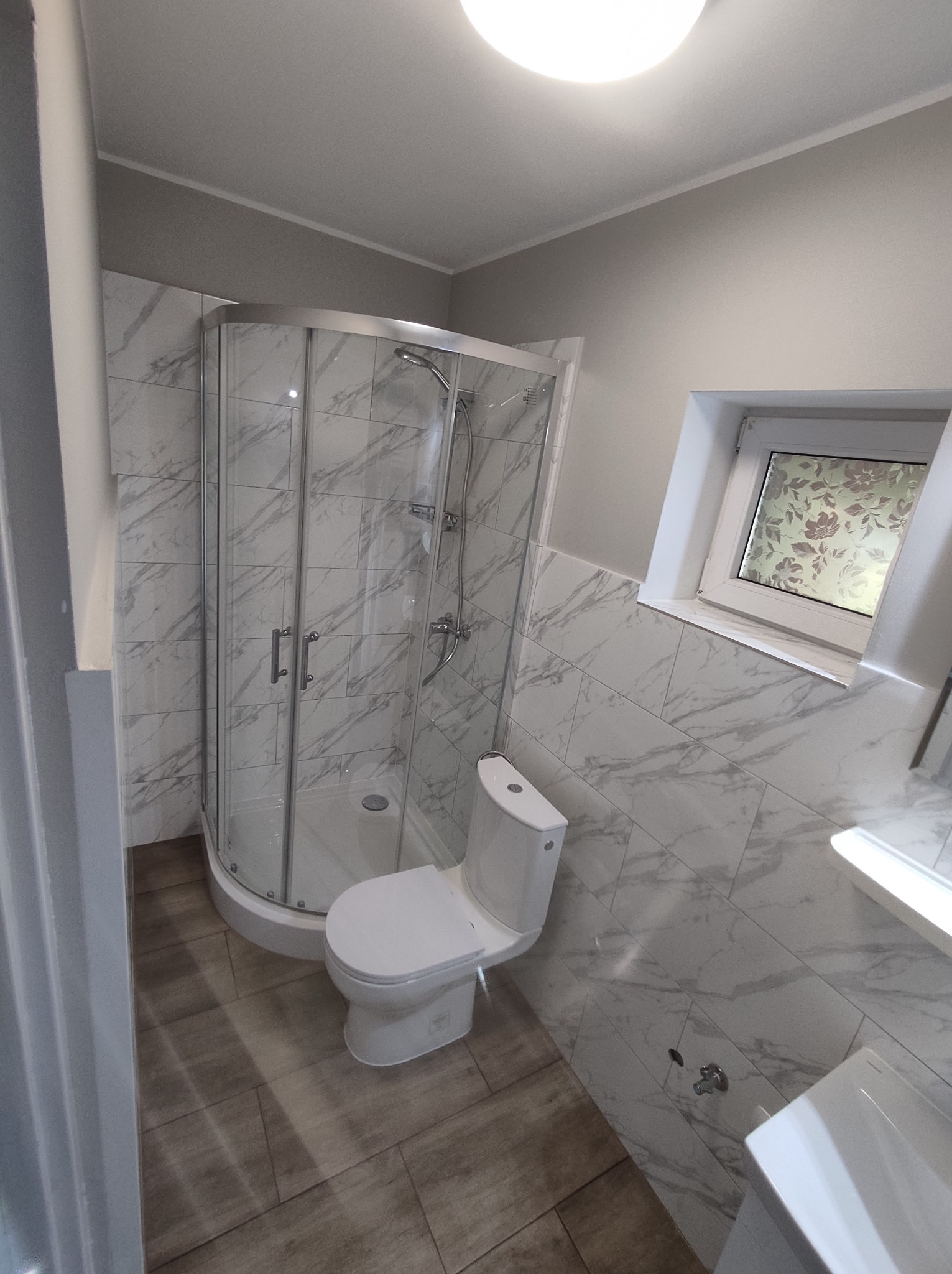 Ukończona łazienka z kabiną prysznicową z zaokrąglonymi drzwiami, toaletą typu kompakt i umywalką, wykończona płytkami imitującymi biały marmur z szarymi żyłami oraz drewnopodobnymi płytkami...