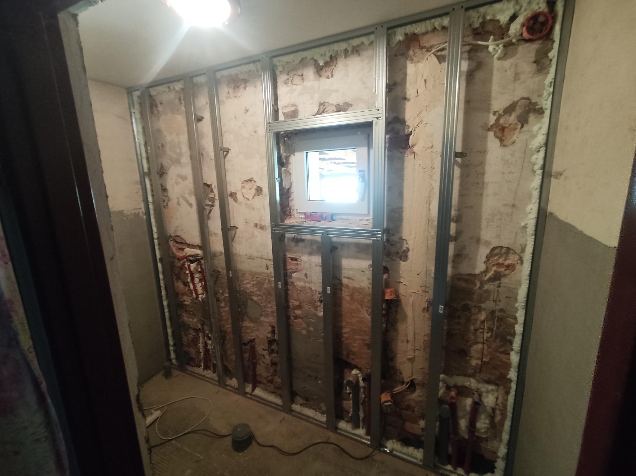 Wnętrze w trakcie prac przygotowawczych do wykończenia ścian, widoczne profile stalowe, nowo wstawione okno i fragmenty starego muru z instalacjami.