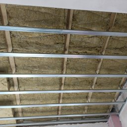 POZIOM JEST V. MIK & A. SPANCHAK S.C. - Sufit poddasza w trakcie ocieplania wełną mineralną, widoczne drewniane belki, metalowe profile i siatka podtrzymująca izolację.