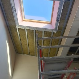 POZIOM JEST V. MIK & A. SPANCHAK S.C. - Montaż okna dachowego: widok z dołu na izolowane poddasze z widocznym oknem dachowym, metalową konstrukcją nośną i fragmentem rusztowania.