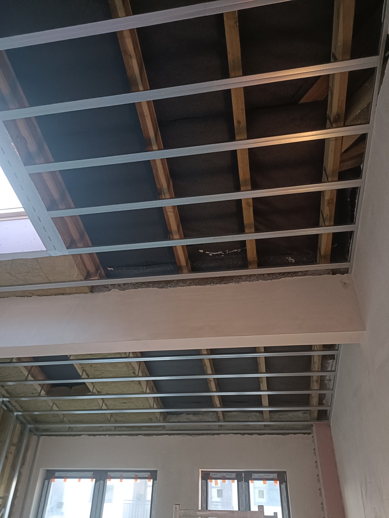 Sufit podwieszany w trakcie montażu: widoczne metalowe profile, izolacja z wełny mineralnej i drewniane elementy konstrukcyjne, częściowo zakryte czarną folią, ściany w stanie surowym.