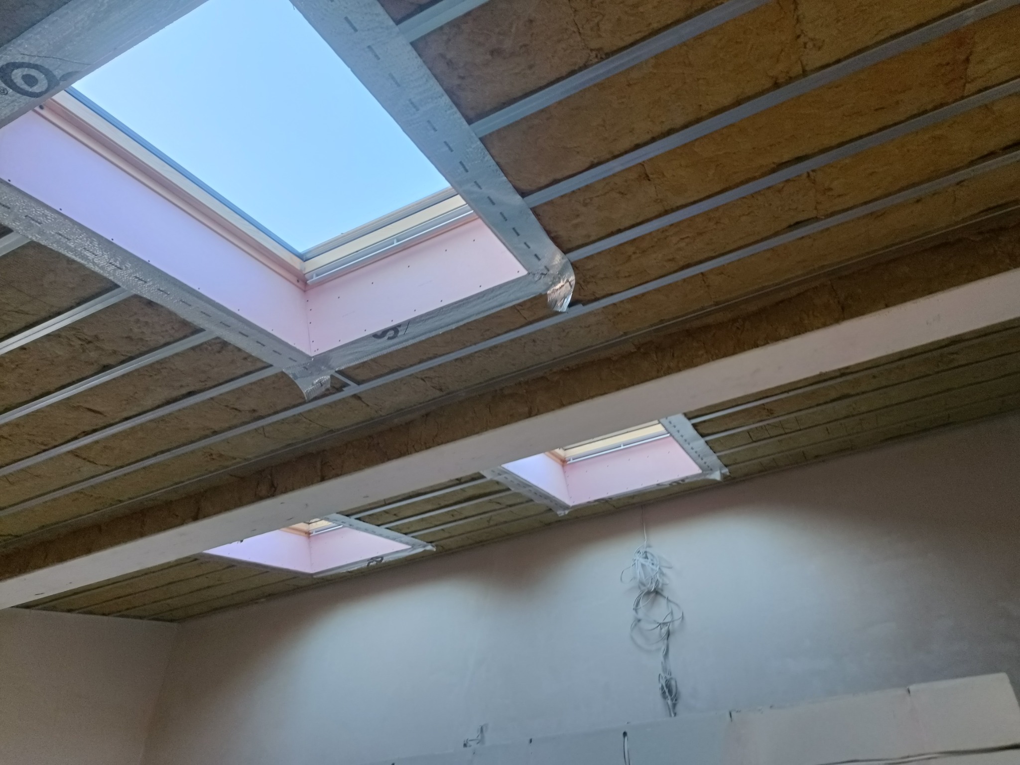 Poddasze z oknami dachowymi w trakcie ocieplania, widoczne wełna mineralna między krokwiami i metalowy stelaż, różowe obramowania okien, wiszące kable.