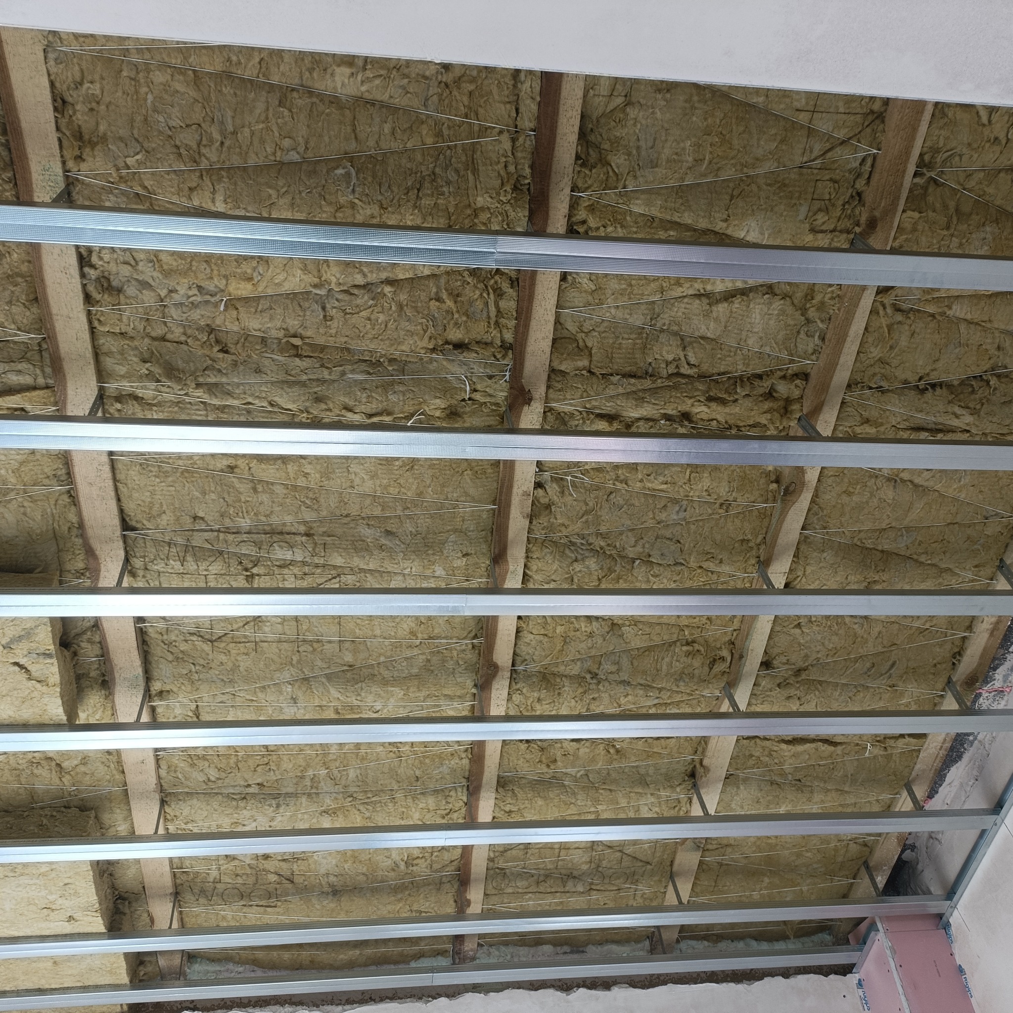 Sufit poddasza w trakcie ocieplania wełną mineralną, widoczne drewniane belki, metalowe profile i siatka podtrzymująca izolację.