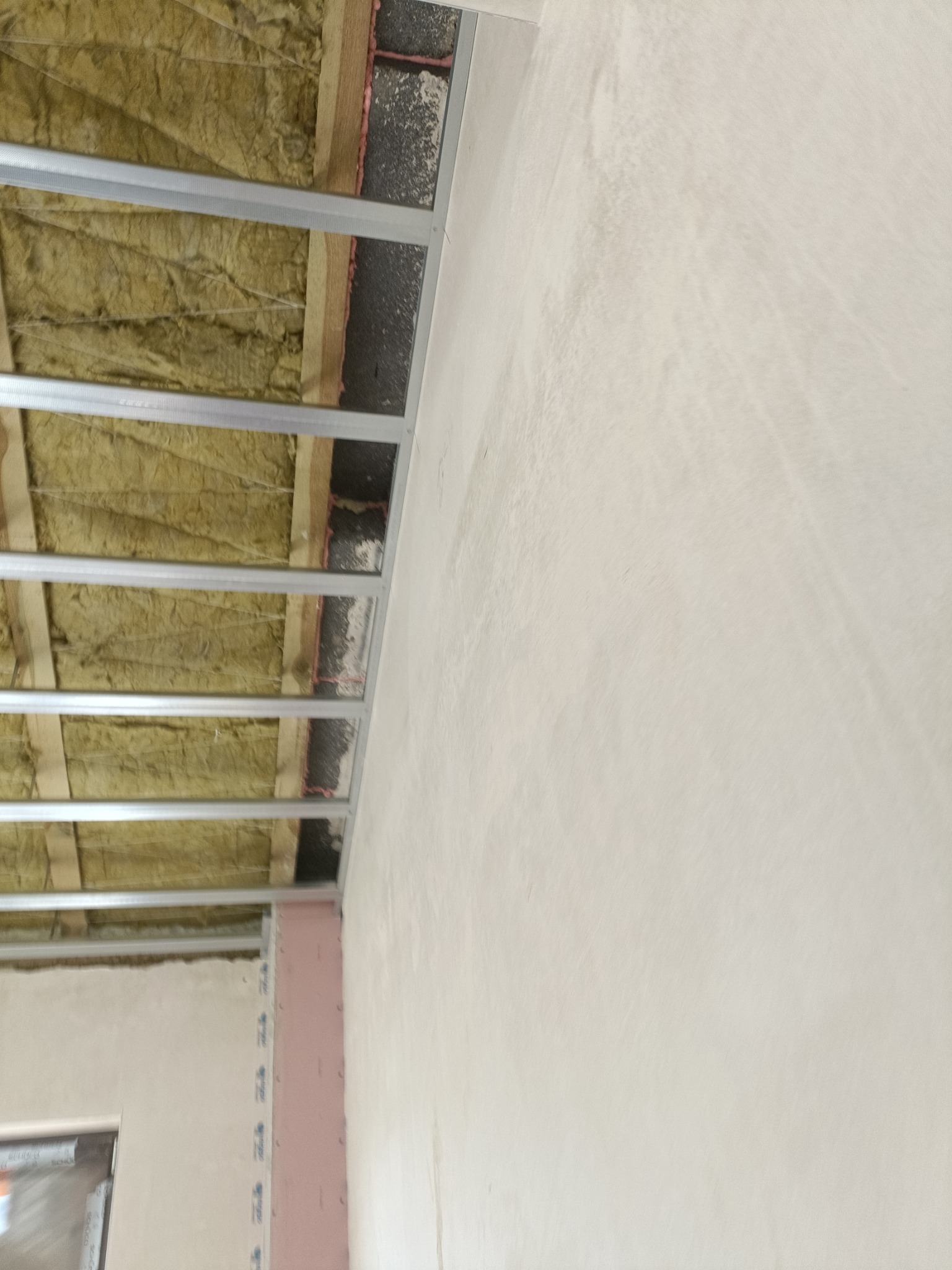 Szkielet sufitu podwieszanego z izolacją z wełny mineralnej, widoczna ściana z surową powierzchnią przed wykończeniem.