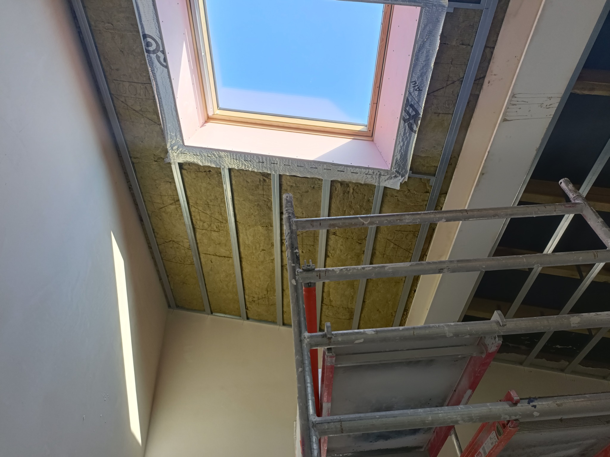 Montaż okna dachowego: widok z dołu na izolowane poddasze z widocznym oknem dachowym, metalową konstrukcją nośną i fragmentem rusztowania.