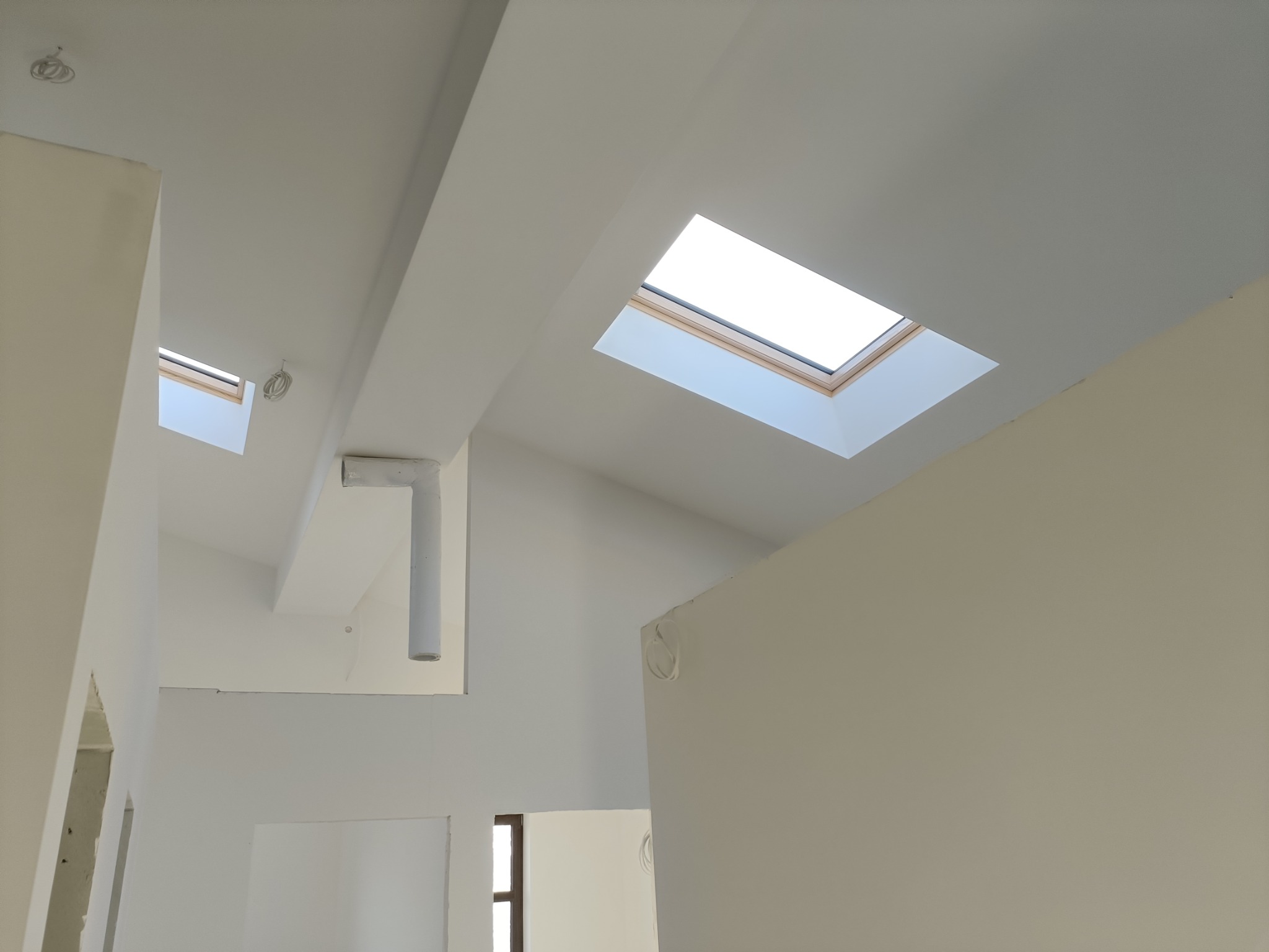 Wnętrze w trakcie prac wykończeniowych: białe ściany i sufit, okna dachowe z drewnianą ramą, widoczne przewody elektryczne na ścianach, surowe wykończenie.