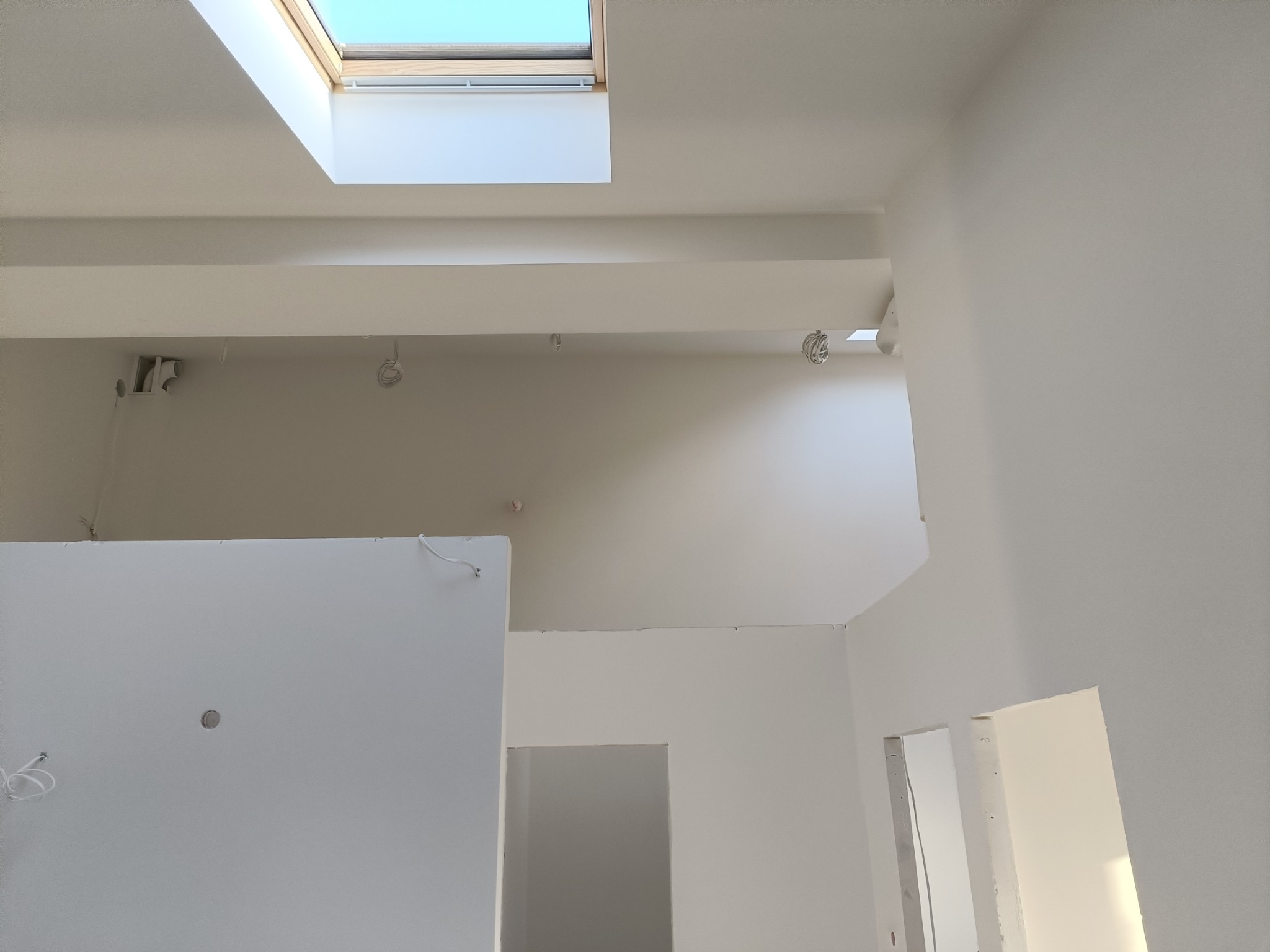Wnętrze w trakcie wykańczania, widoczne ściany pomalowane na biało, sufit z oknem dachowym,  przygotowane punkty oświetleniowe i fragment ściany z płyty karton gips.