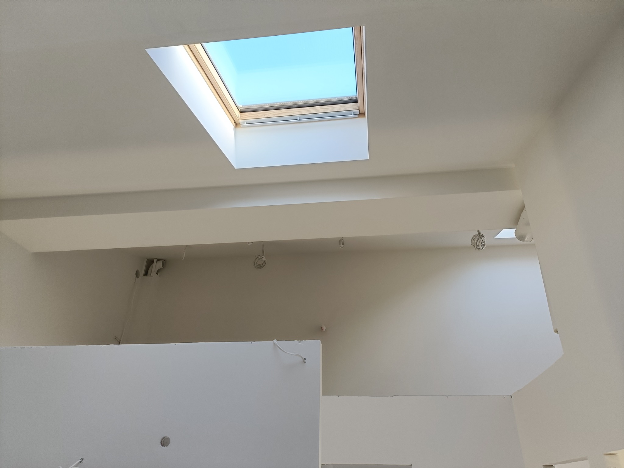 Wnętrze w trakcie prac, widok na sufit z oknem dachowym, widoczne niedokończone ściany działowe z płyt kartonowo-gipsowych i instalacje elektryczne.