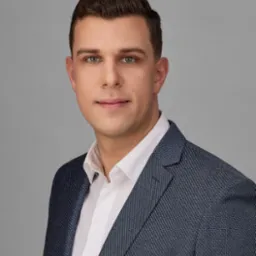 Portret młodego mężczyzny w niebieskiej marynarce i białej koszuli, z białą poszetką w kieszonce, na jednolitym szarym tle.