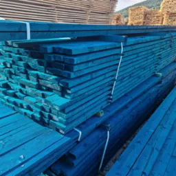 Stosy tarcicy budowlanej pomalowanej na intensywny niebieski kolor, zabezpieczone białymi taśmami, ułożone na zewnątrz z widocznymi górami w tle.