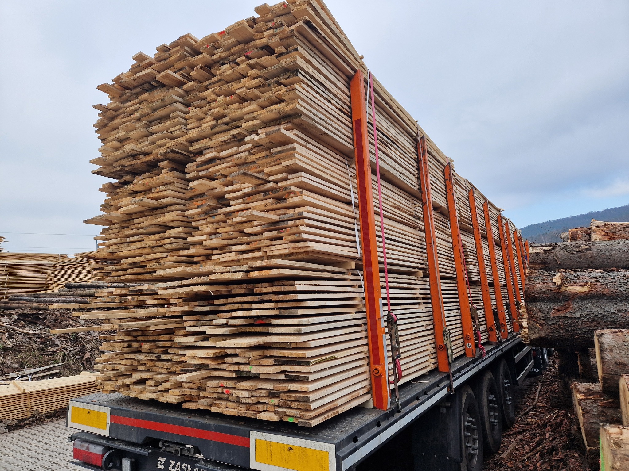 Ciężarówka załadowana stertą świeżo przetartej tarcicy budowlanej, zabezpieczonej pomarańczowymi pasami transportowymi, na tle pochmurnego nieba i składowanych bali drewna.