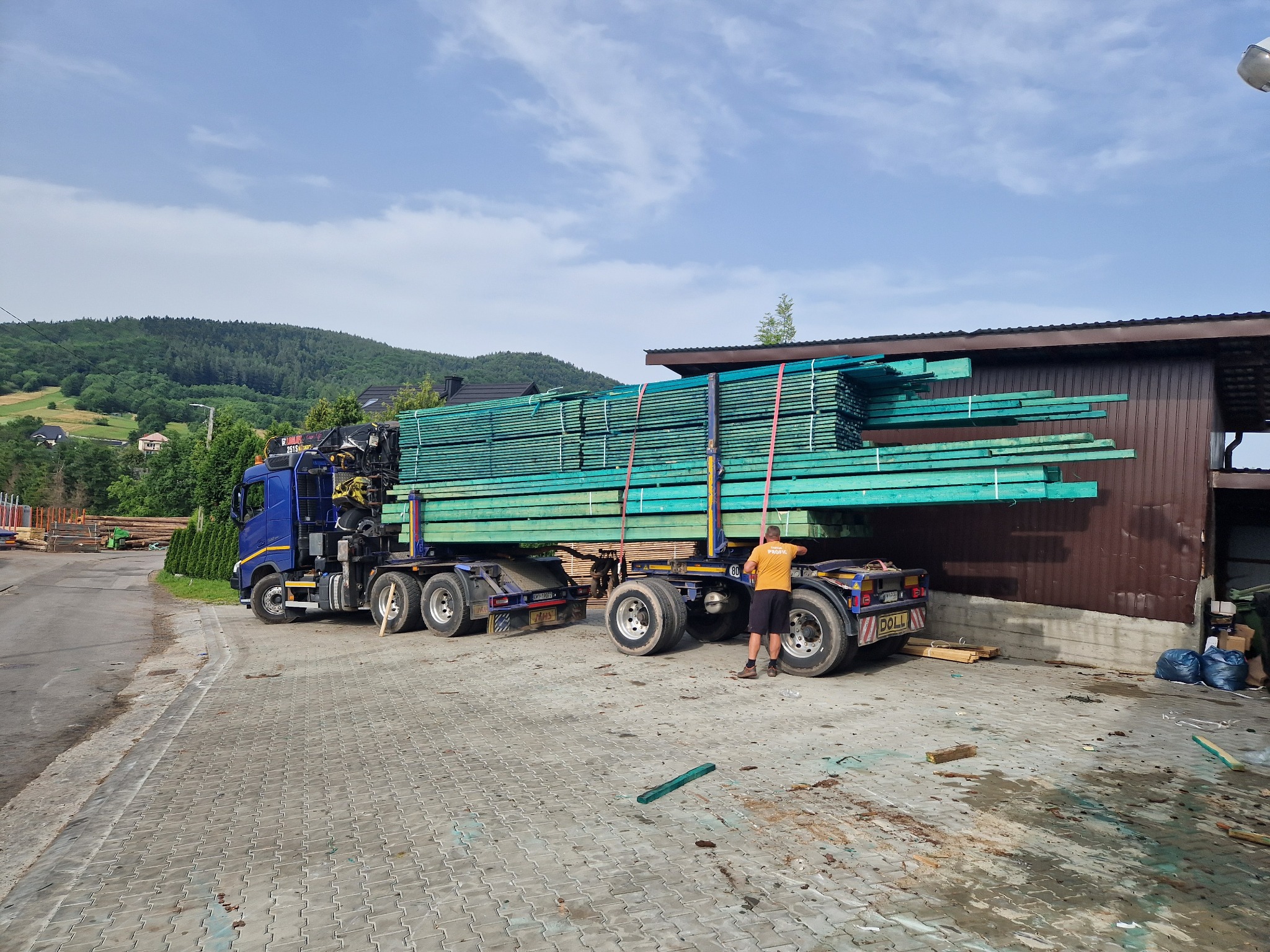 Niebieska ciężarówka z naczepą załadowana świeżo pomalowaną na zielono tarcicą budowlaną, mężczyzna w pomarańczowej koszulce zabezpiecza ładunek, w tle budynek magazynowy i zalesione wzgórza.