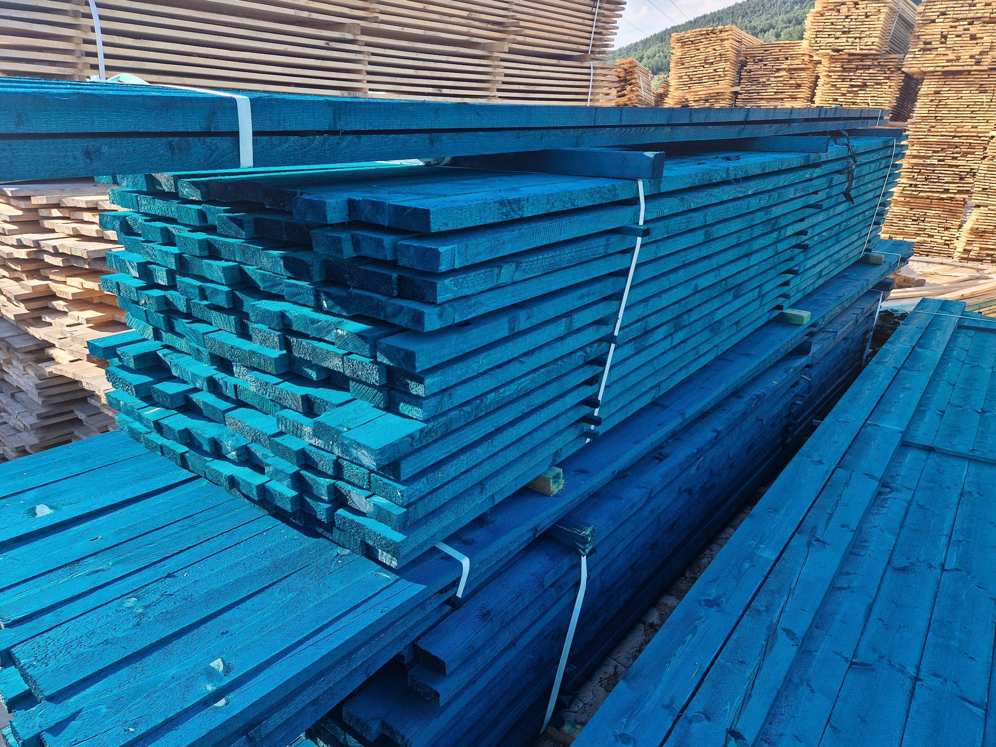 Stosy tarcicy budowlanej pomalowanej na intensywny niebieski kolor, zabezpieczone białymi taśmami, ułożone na zewnątrz z widocznymi górami w tle.