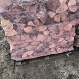 Ułożone na palecie worki z pociętym drewnem kominkowym, zabezpieczone różową siatką, na ubłoconym podłożu.