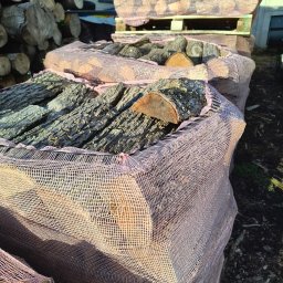 FIRMA HANDLOWO-USŁUGOWA KRZYSZTOF EDEL - Ułożone worki z pociętymi kawałkami drewna, częściowo zasłonięte siatką w kolorze różowym, widoczne pęknięcia na korze