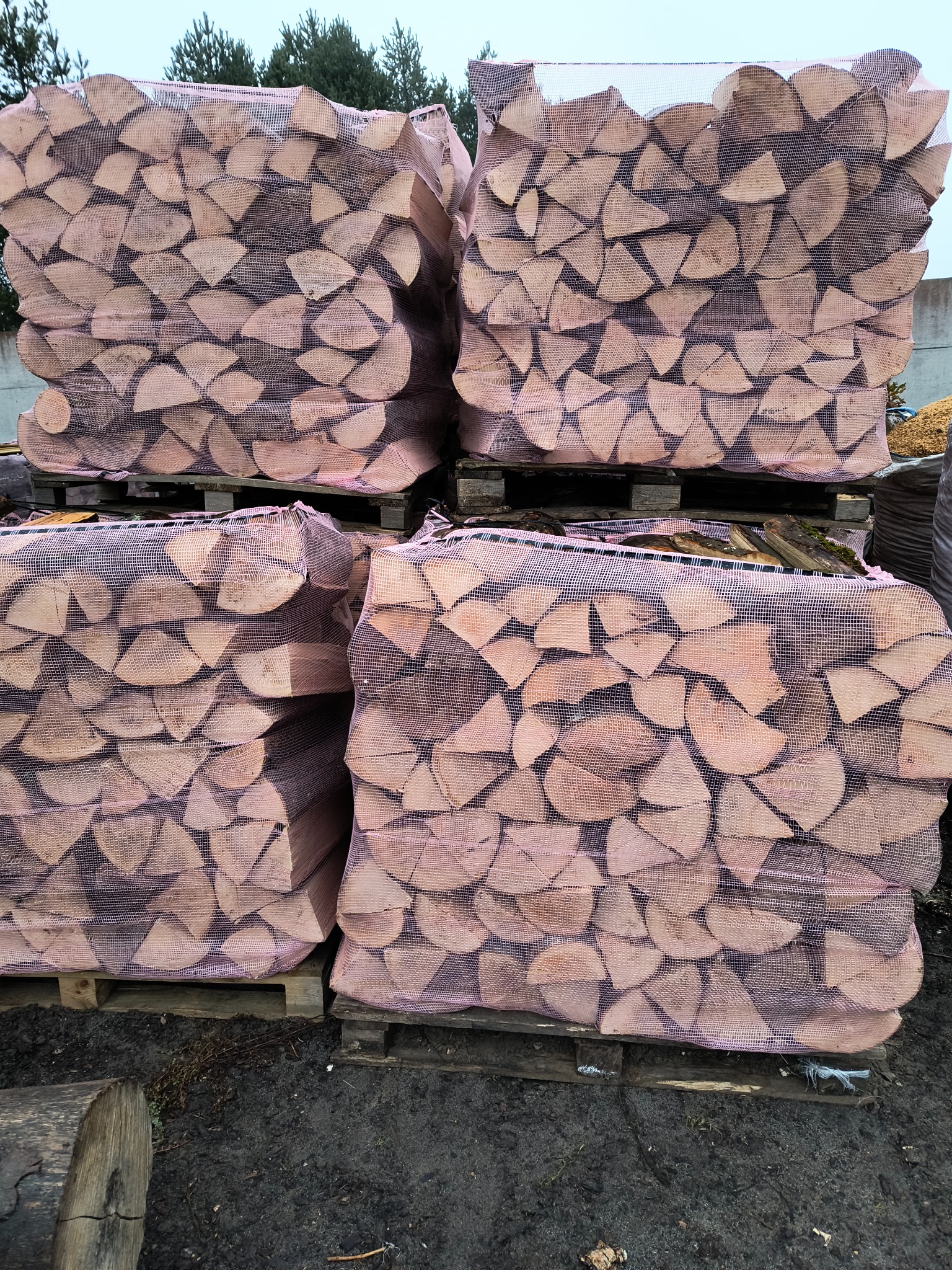 Ułożone na paletach worki z porąbanym drewnem, zabezpieczone różową siatką, widoczne na tle drzew.