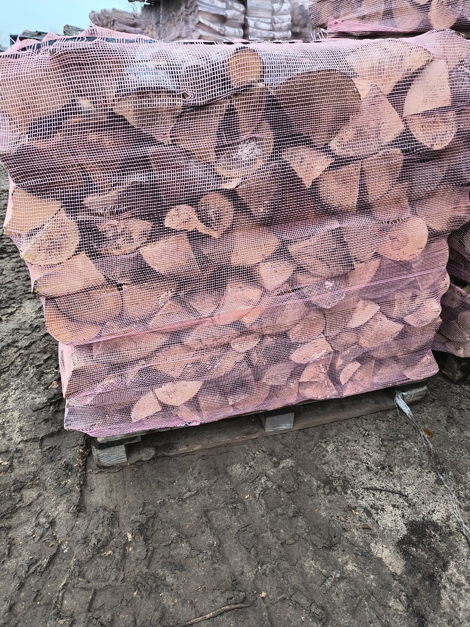 Ułożone na palecie worki z pociętym drewnem kominkowym, zabezpieczone różową siatką, na ubłoconym podłożu.