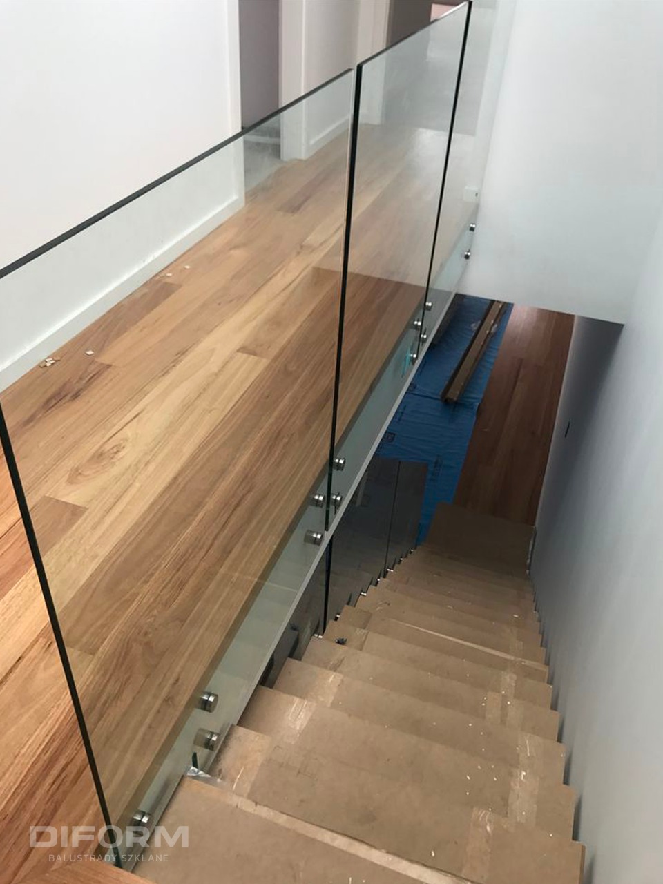 Szklana balustrada na schodach z drewnianymi stopniami, widok z góry, mocowana punktowo, z widocznym zabezpieczeniem podłogi.