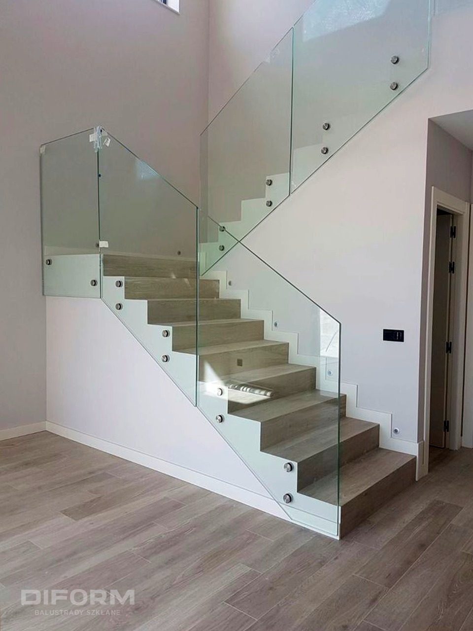 Nowoczesne schody z drewnianymi stopniami i przezroczystą szklaną balustradą, mocowaną punktowo, widok z dołu na jasne wnętrze.