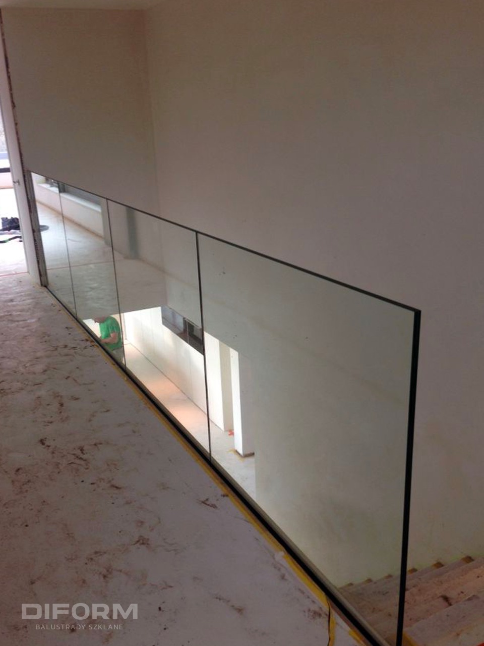 Wnętrze w trakcie remontu z zamontowaną szklaną balustradą z czarnymi krawędziami, widok z góry na podłogę zabezpieczoną folią.