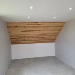 Poddasze z drewnianą boazerią na skosie, wykończone szarą farbą na ścianach i suficie, z wbudowanymi punktowymi lampami sufitowymi.