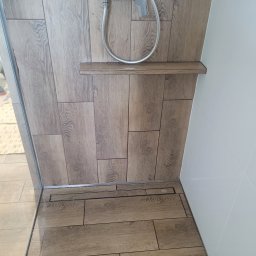 DudziakFach - Wykończona kabina prysznicowa z odpływem liniowym i drewnopodobnymi płytkami na ścianach. Półka w kabinie i biała ściana po prawej stronie.