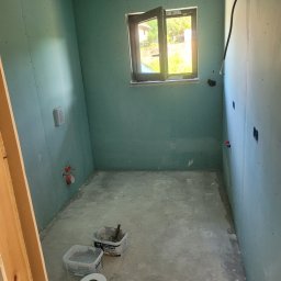 DudziakFach - Łazienka w trakcie przygotowań do wykończenia: ściany obłożone płytami gipsowo-kartonowymi, widoczne puszki elektryczne, otwarte okno i narzędzia na podłodze.