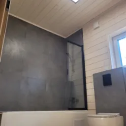 Nowoczesna łazienka z szarą glazurą, drewnianym sufitem i kabiną prysznicową z czarnymi ramami. Uwagę przyciąga wisząca toaleta i mała klapka rewizyjna.