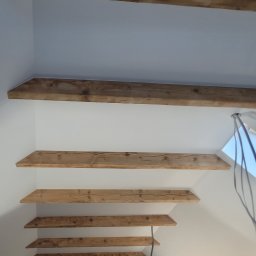 DudziakFach - Wykończone drewniane belki stropowe na białym poddaszu, widok z dołu. Element dekoracyjny wnętrza z widocznymi słojami drewna i fragmentem okna dachowego.