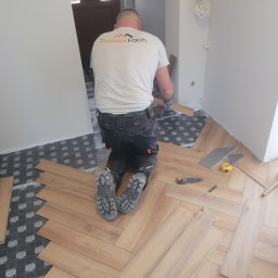 DudziakFach - Układanie paneli podłogowych w jodełkę. Widoczny pracownik na kolanach, narzędzia: nóż, miarka. Częściowo ułożona podłoga na podkładzie. Jasne wnętrze.