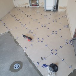 DudziakFach - Układanie jasnych płytek podłogowych z użyciem systemu poziomowania. Widoczny fragment surowej posadzki, narzędzia glazurnicze i instalacja CO w tle.