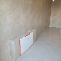 DudziakFach - Wykończona łazienka z beżowymi płytkami imitującymi marmur na ścianach i podłodze. Widoczna zabudowa z drzwiczkami rewizyjnymi, zabezpieczona pomarańczową taśmą.