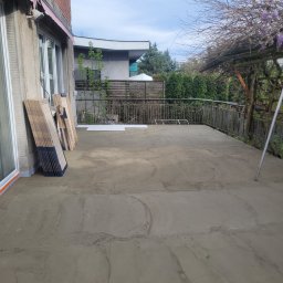DudziakFach - Przygotowany betonowy podkład na balkonie, gotowy do położenia paneli. Paczki paneli oparte o ścianę. Widok na zewnątrz z metalową barierką.