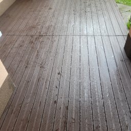 DudziakFach - Ułożona, brązowa deska tarasowa z widocznymi kroplami wody, przylegająca do ściany budynku. Widoczne połączenia desek i fragment trawnika w tle.