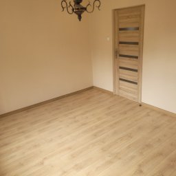 DudziakFach - Jasny pokój z nowo ułożonymi panelami podłogowymi w odcieniu dębu, beżowymi ścianami, nowoczesnymi drzwiami z ciemnymi wstawkami i klasycznym żyrandolem.