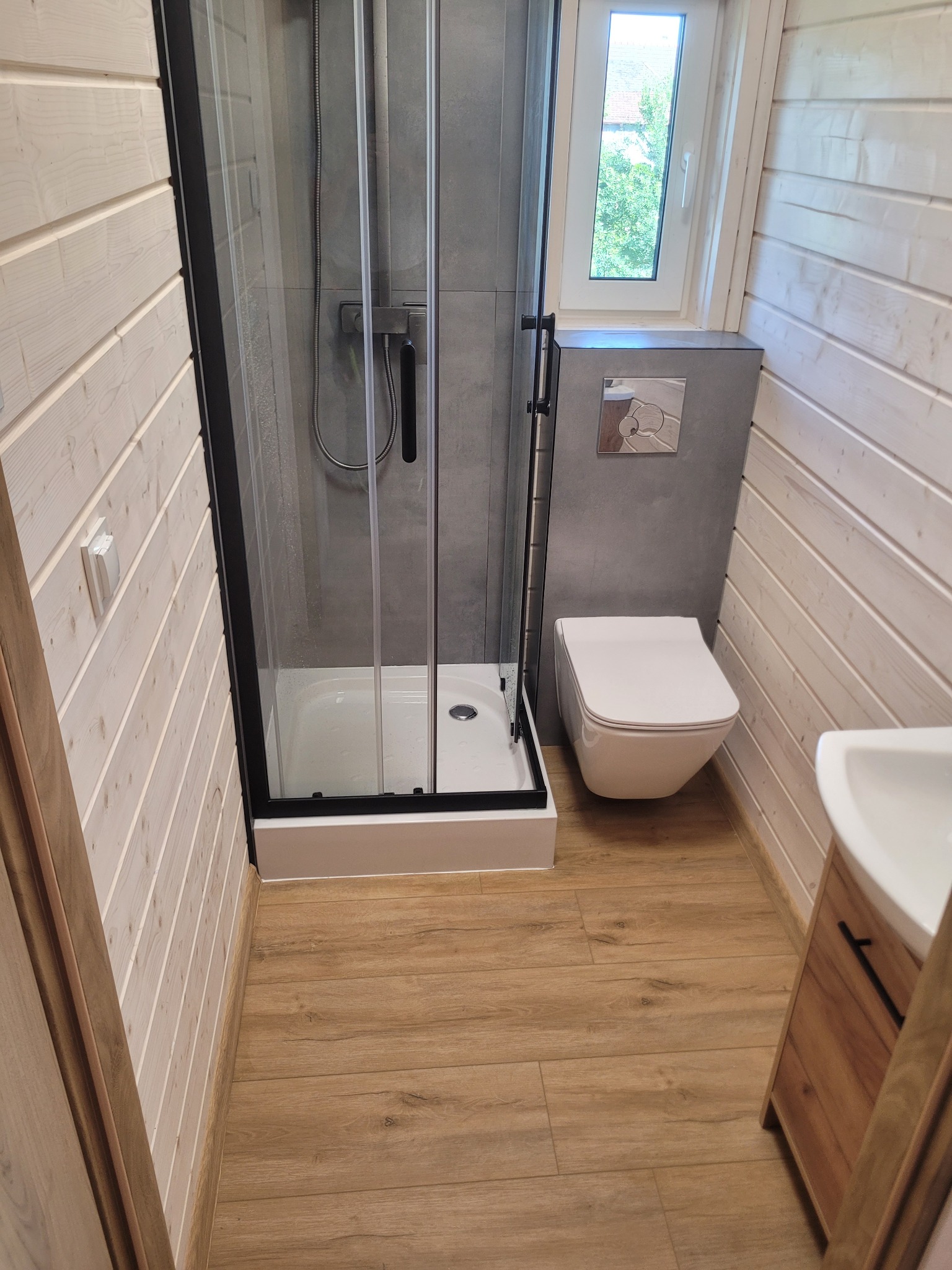 Wykończona łazienka w stylu skandynawskim: kabina prysznicowa z czarną ramą, podwieszana toaleta, drewniane panele ścienne i podłoga imitująca drewno.