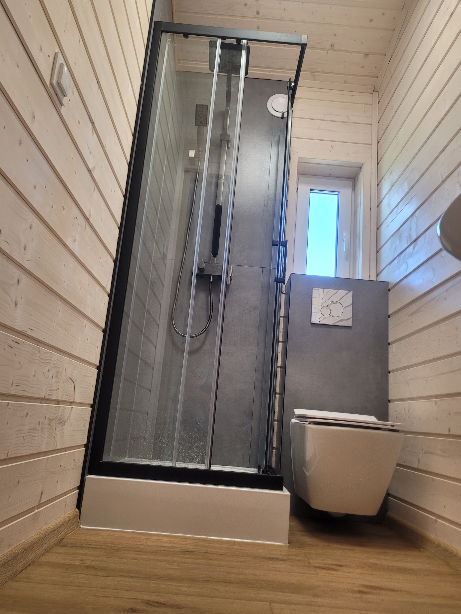 Nowoczesna łazienka z kabiną prysznicową typu walk-in, drewnianą boazerią i podwieszaną toaletą. Minimalistyczne wnętrze z szarymi akcentami i jasnym oświetleniem.