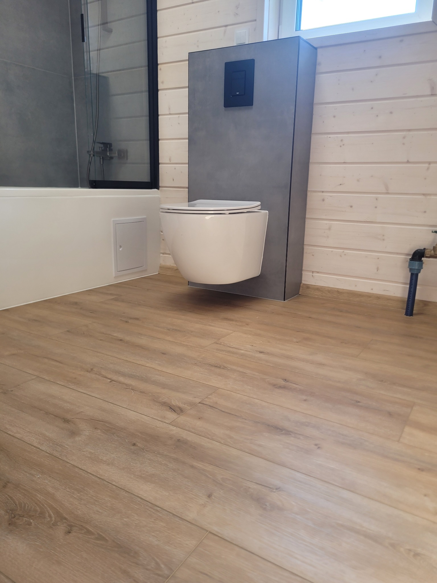 Nowoczesna łazienka w drewnie: podwieszana toaleta z szarą zabudową, drewniana podłoga, biała wanna z czarną szybą prysznicową i okno w tle.