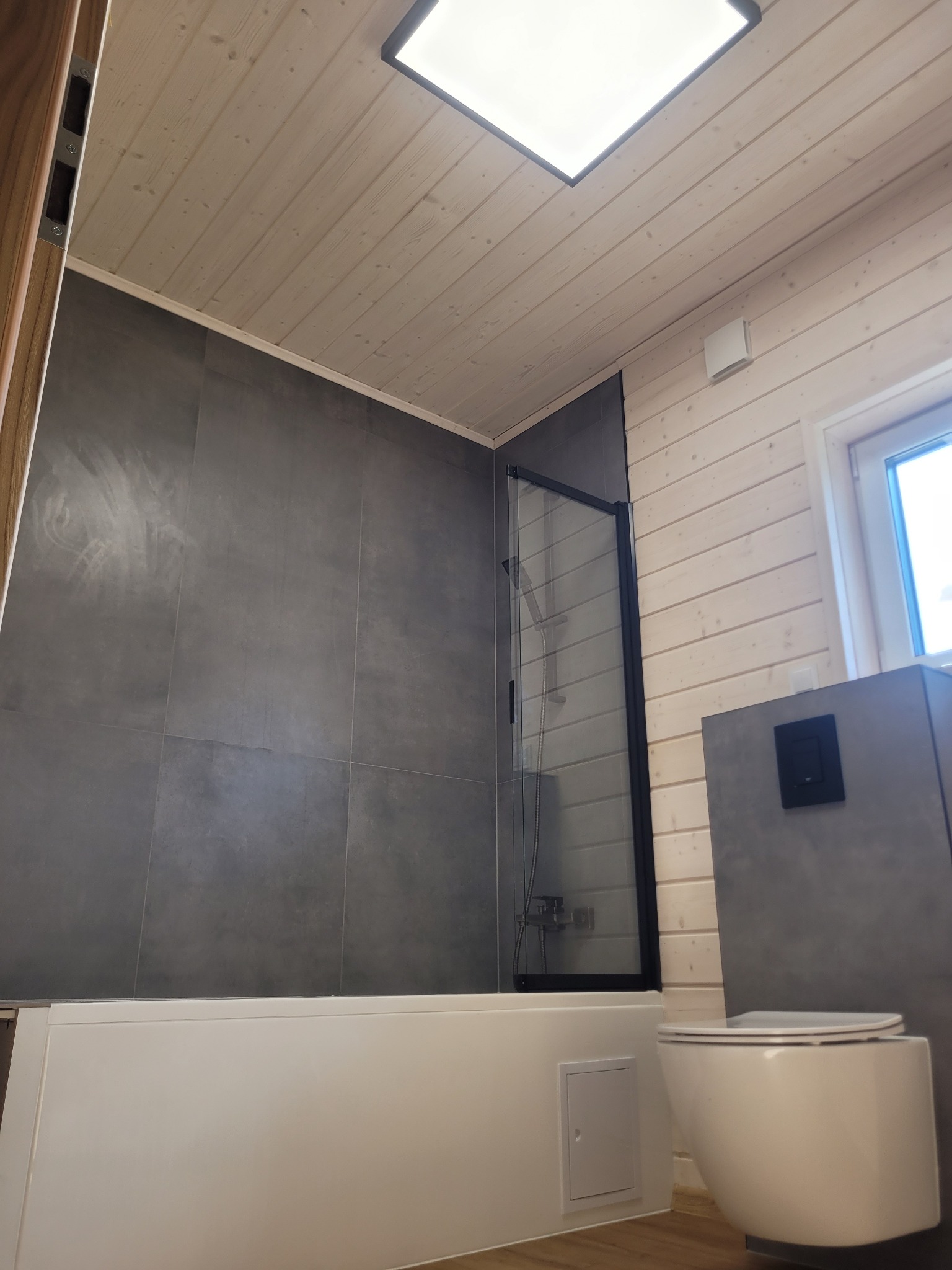Nowoczesna łazienka z szarą glazurą, drewnianym sufitem i kabiną prysznicową z czarnymi ramami. Uwagę przyciąga wisząca toaleta i mała klapka rewizyjna.