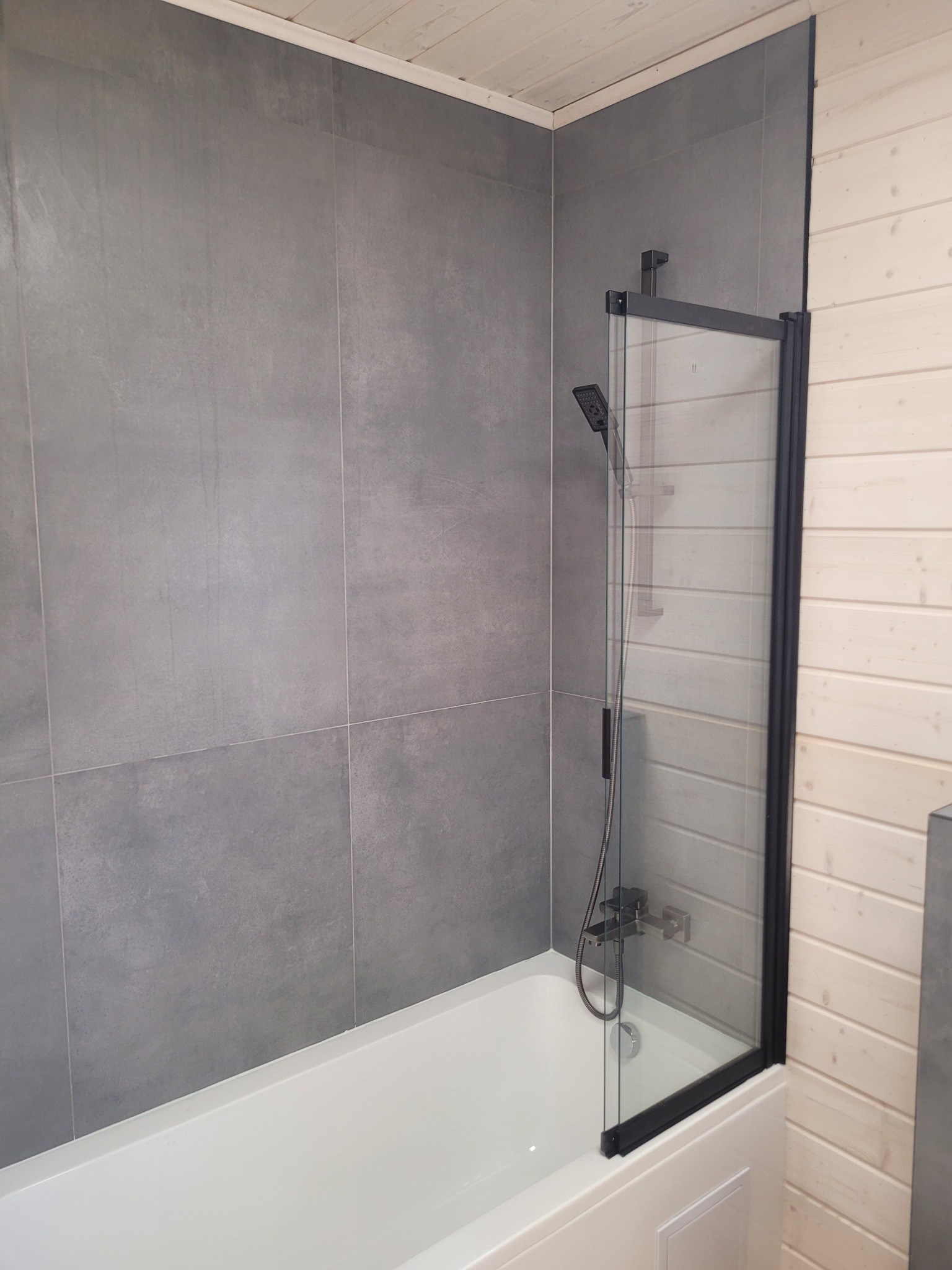 Nowoczesna łazienka: biała wanna z czarnym parawanem nawannowym, szare płytki ścienne i jasne drewniane panele. Minimalistyczny design z akcentem na funkcjonalność.