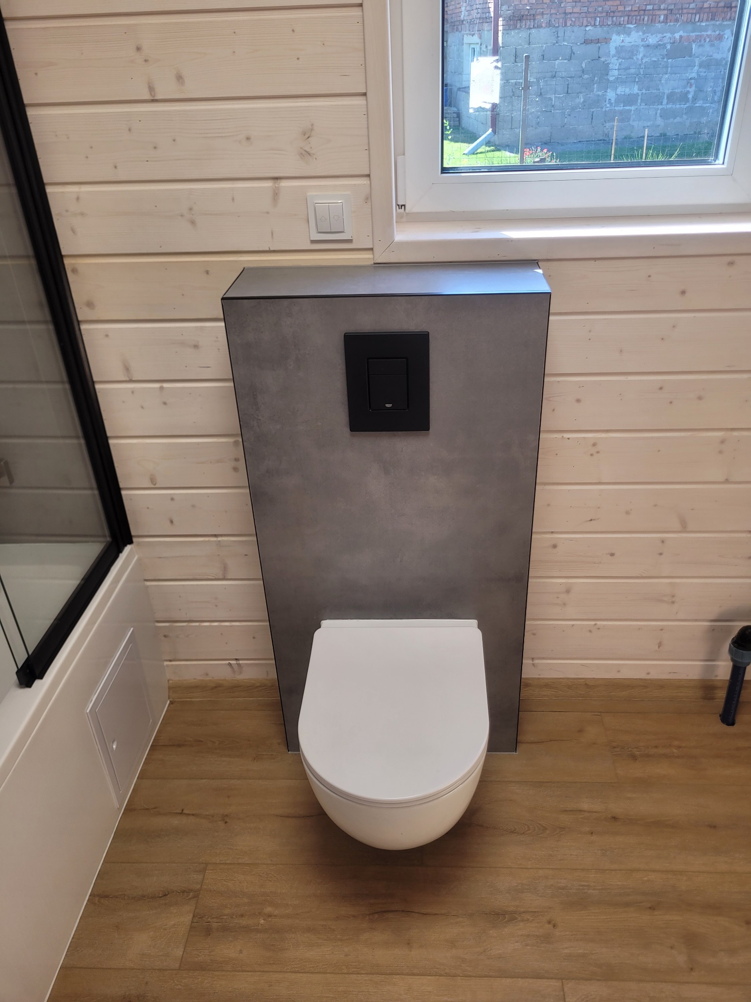 Nowoczesna toaleta podwieszana z szarą zabudową na tle jasnych paneli ściennych i drewnopodobnej podłogi. Widoczne okno i fragment kabiny prysznicowej.