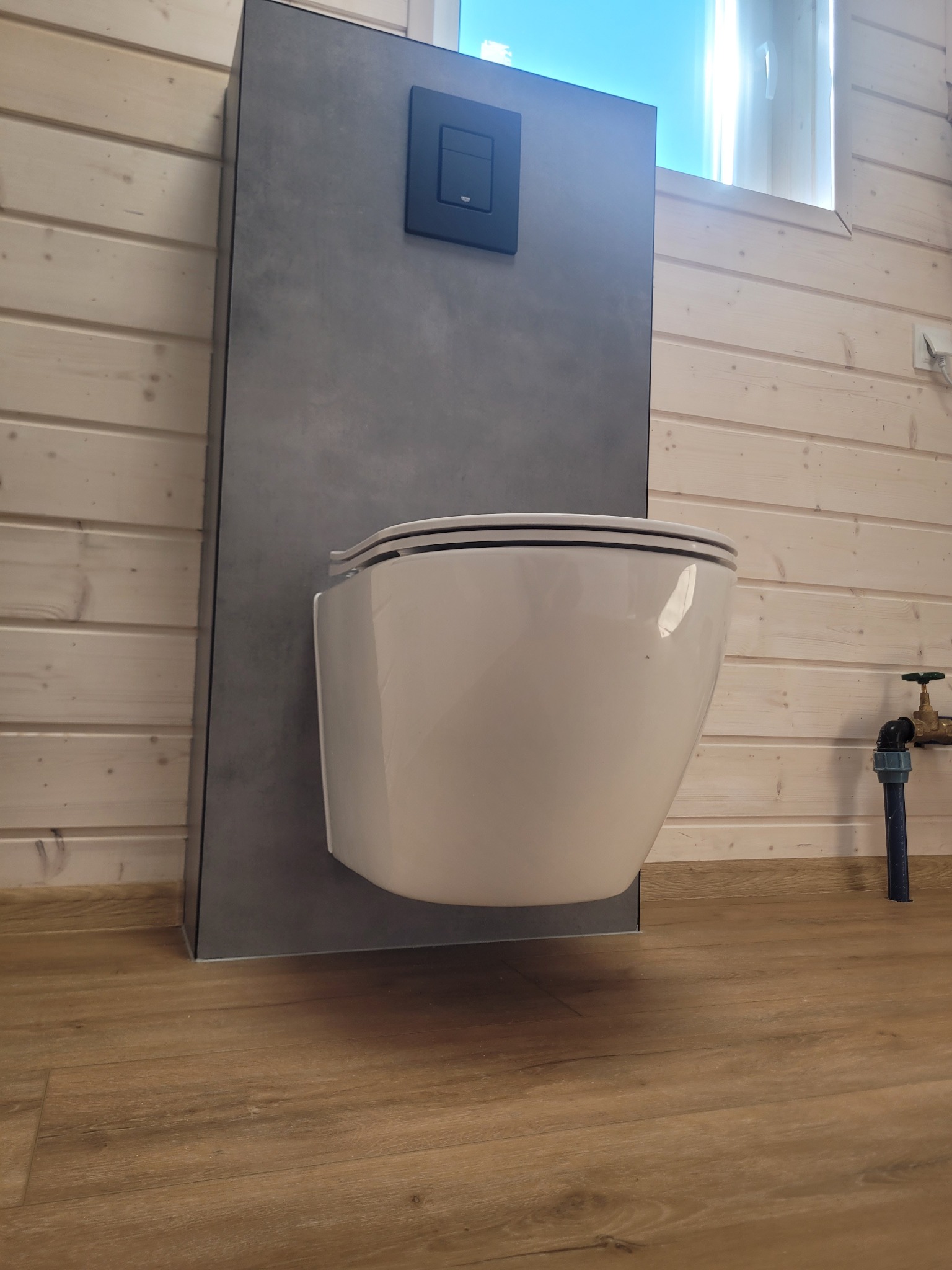 Nowoczesna toaleta podwieszana z szarą zabudową i czarnym przyciskiem spłukującym na tle drewnianej boazerii i paneli podłogowych. Minimalistyczny design.